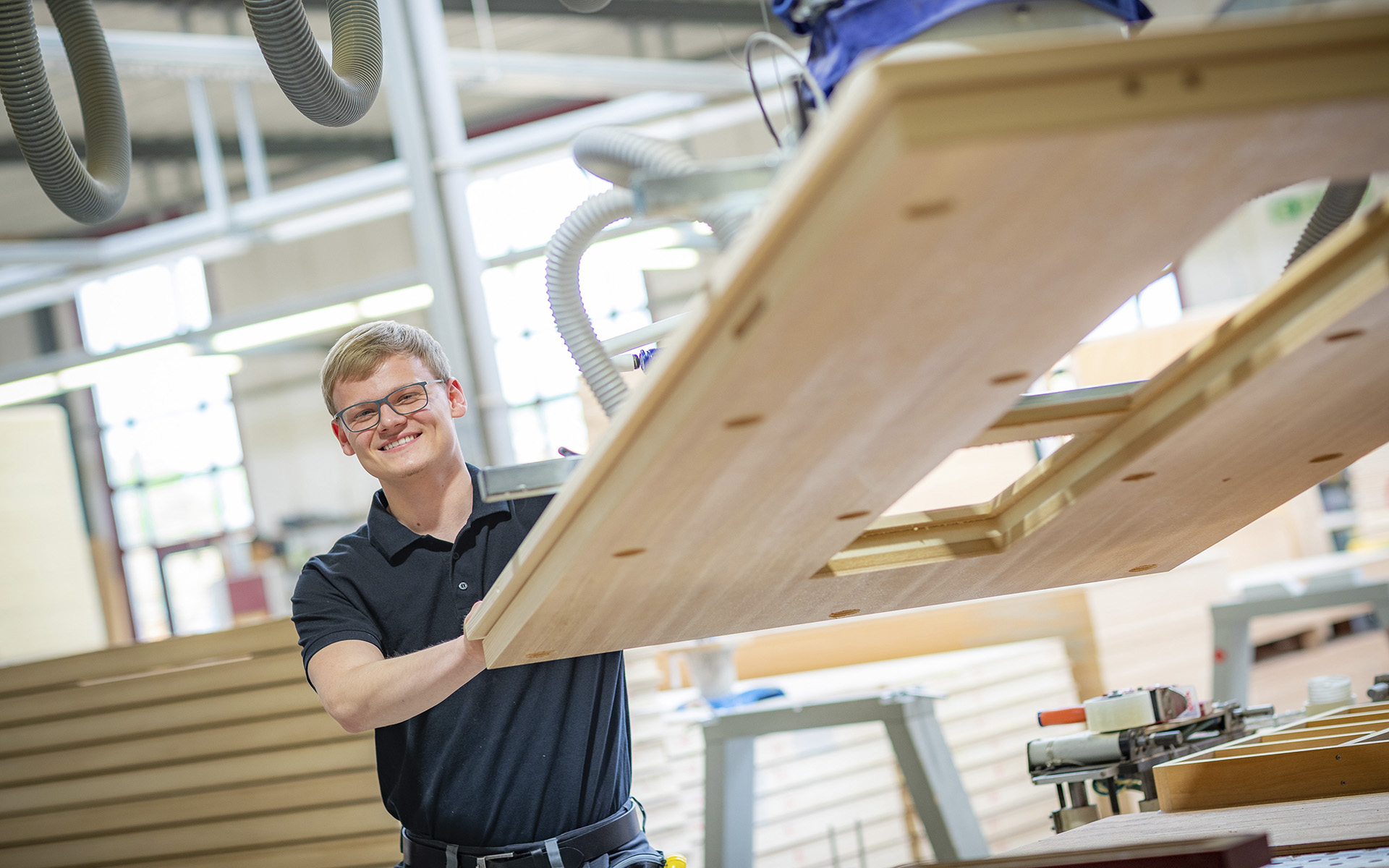 Ein lächelnder Mann in schwarzem Polohemd arbeitet in einer Holzwerkstatt.