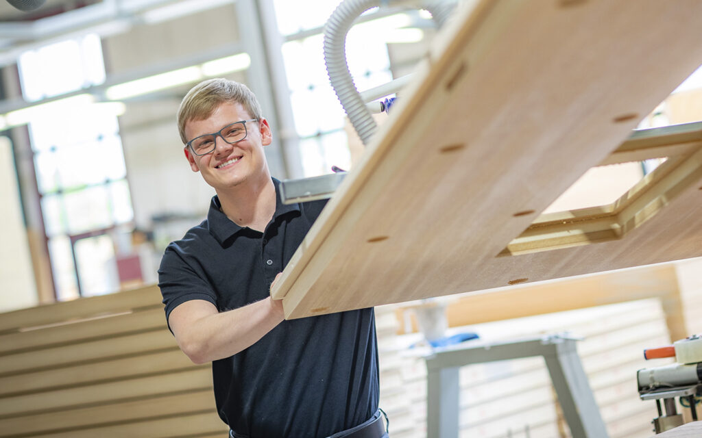 Ein lächelnder Mann in schwarzem Polohemd arbeitet an einem Holztisch.