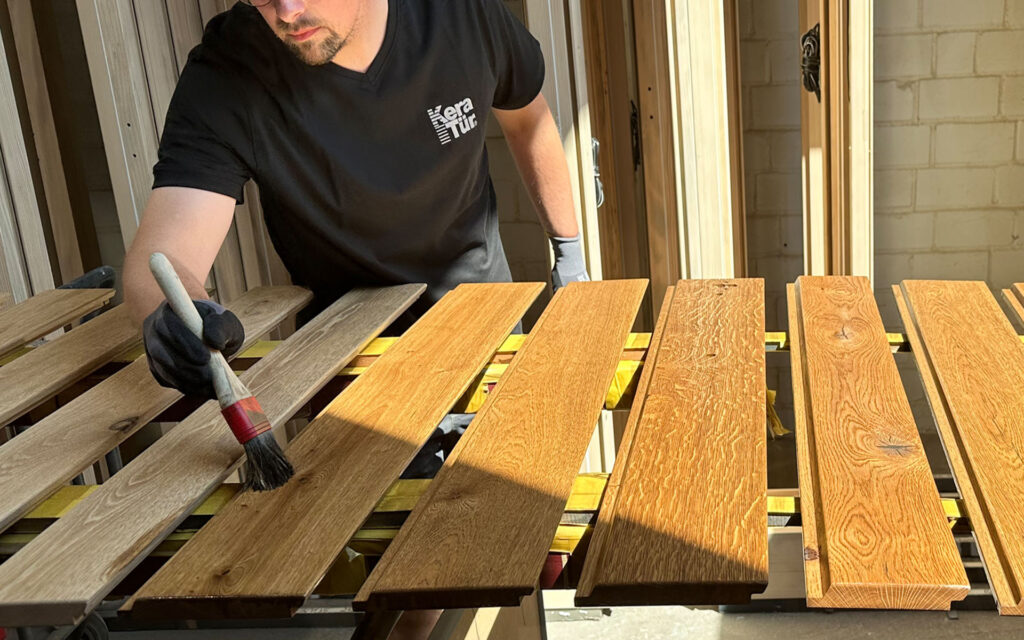 Ein Handwerker streicht Holzbretter in sonnigem Licht mit konzentriertem Ausdruck.