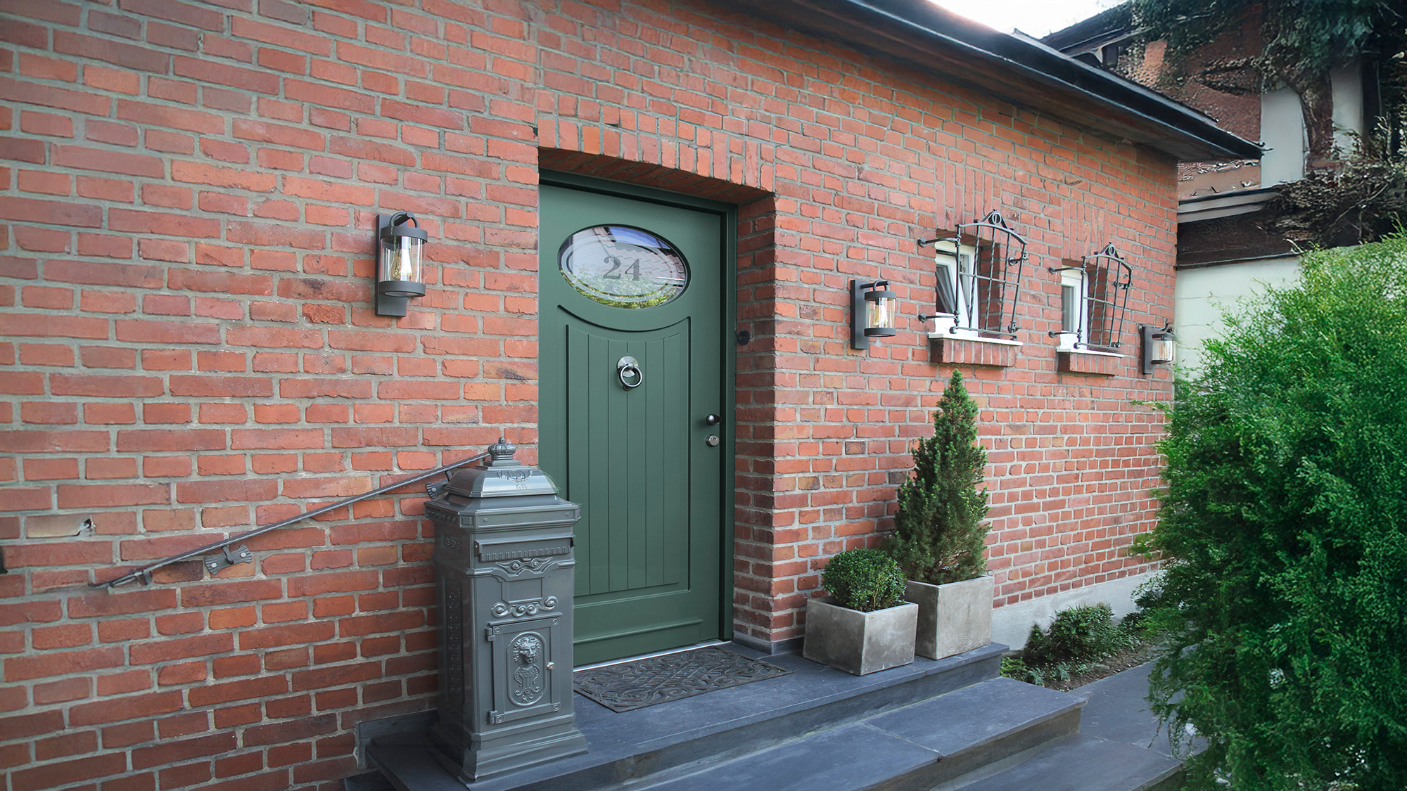 Rote Backsteinfassade mit gruenem Tuer, Briefkasten und Pflanzen, ruhig